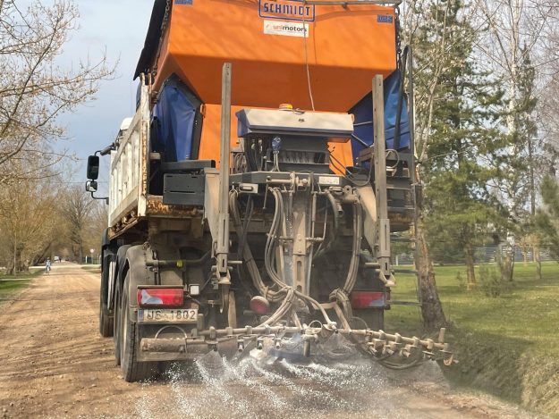 2021_04_20_atputekl1_kulk Kravas automašīna uz grants ceļa veic pretputekļu apstrādi, kaisot reaģentu no speciālas oranžas iekārtas. Fonā redzama ceļmala ar kokiem pavasara dienā.