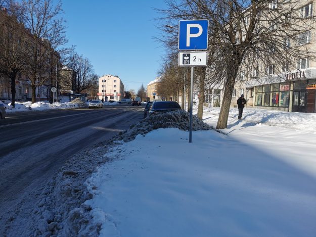 Matera_iela_2h Pilsētas iela ziemā ar uzstādītu autostāvvietas zīmi, kas atļauj stāvēšanu līdz 2 stundām. Ceļa malā novietotas automašīnas, redzami sniega vaļņi, koki un dzīvojamās ēkas.
