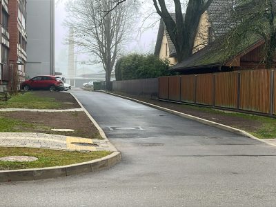 Asfaltēta piebraucamā iela starp daudzstāvu dzīvojamo ēku un privātmāju teritoriju ar koka žogu. Ceļa malā novietota automašīna, redzams ietves segums un pagalma piebrauktuve.