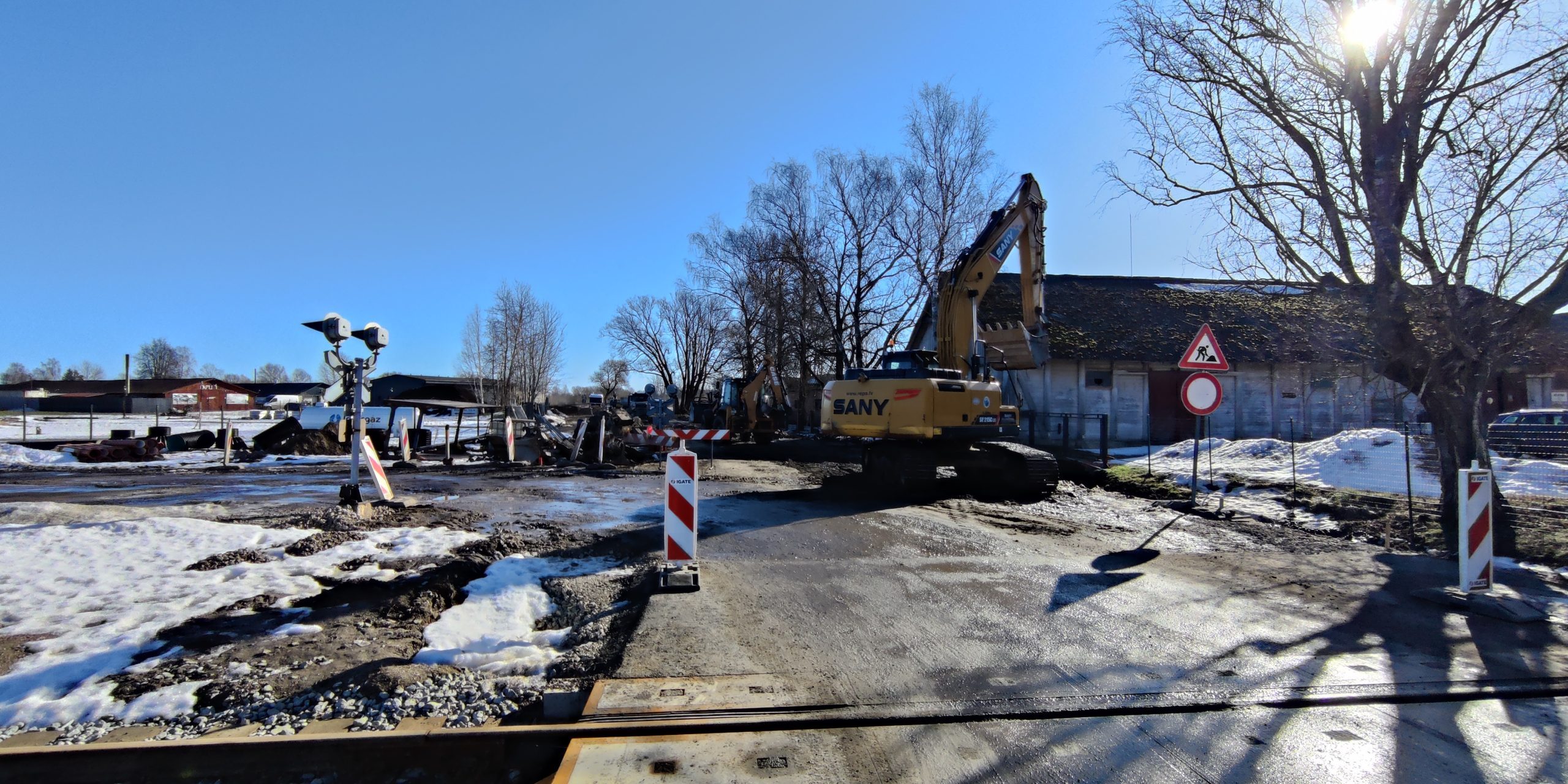 Būvdarbu norise Dzirnavu ielā. Ceļa būvdarbi pie dzelzceļa pārbrauktuves – uz brauktuves redzams ekskavators, uzstādītas pagaidu satiksmes organizācijas zīmes un norobežojošie stabiņi. Apkārt redzams būvdarbu laukums ar tehniku, sniegu un ēkām fonā.