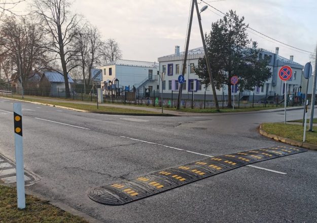 Lacplesa_Iecavas_k_atrumv_PUBL-1024×720 Melns gumijas ātrumvalnis ar dzelteniem atstarojošiem elementiem uz pilsētas ielas pie krustojuma, fonā redzama sabiedriskā ēka, ceļa zīmes un ietve.