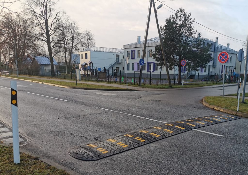 Melns gumijas ātrumvalnis ar dzelteniem atstarojošiem elementiem uz pilsētas ielas pie krustojuma, fonā redzama sabiedriskā ēka, ceļa zīmes un ietve.