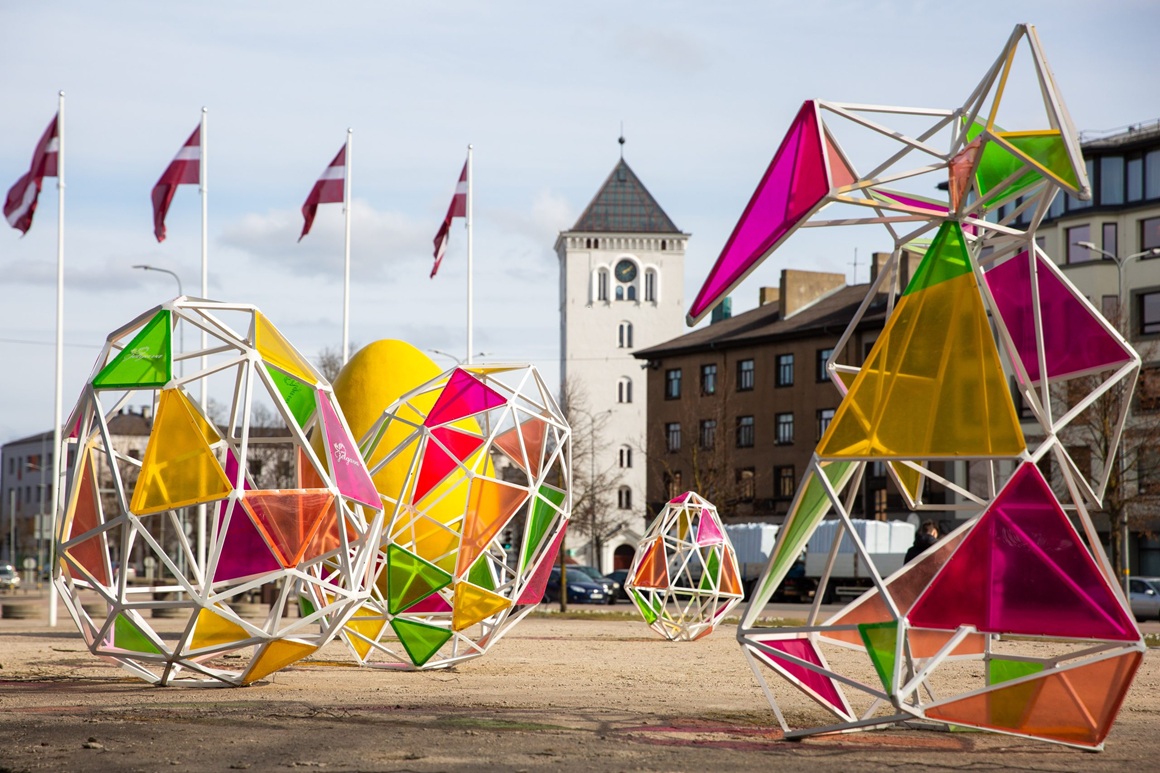 Lieldienu dekorācijas Hercoga Jēkaba laukumā – krāsainas, ģeometriskas, dažādu formu konstrukcijas ar caurspīdīgiem, spilgtiem paneļiem. Fonā redzamas ēkas un vairāki karogi un Svētās Trīsvienības baznīcas tornis.
