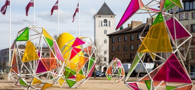 Lieldienu-dekori_small Lieldienu dekorācijas Hercoga Jēkaba laukumā – krāsainas, ģeometriskas, dažādu formu konstrukcijas ar caurspīdīgiem, spilgtiem paneļiem. Fonā redzamas ēkas un vairāki karogi un Svētās Trīsvienības baznīcas tornis.