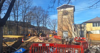 WhatsApp Image 2026-03-06 at 11.37.01 Ceļa būvdarbi pilsētas ielā – redzama izrakta tranšeja, norobežojumi ar sarkanām drošības barjerām, būvtehnika un strādnieki darba procesā. Fonā atrodas ķieģeļu ēka un dzīvojamās mājas, bet priekšplānā redzamas ceļa zīmes, būvmateriāli un izrakta zeme.