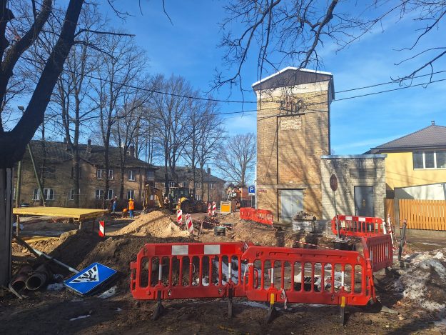 WhatsApp Image 2026-03-06 at 11.37.01 Ceļa būvdarbi pilsētas ielā – redzama izrakta tranšeja, norobežojumi ar sarkanām drošības barjerām, būvtehnika un strādnieki darba procesā. Fonā atrodas ķieģeļu ēka un dzīvojamās mājas, bet priekšplānā redzamas ceļa zīmes, būvmateriāli un izrakta zeme.