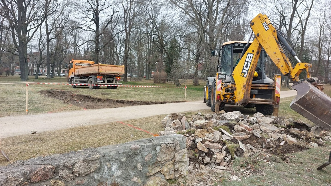 Alunāna parka teritorijā notiek būvdarbi. Ekskavators ar kausu demontē un pārvieto vecas akmens konstrukcijas, blakus izrakta tranšeja gar celiņu un aiz ekskavatora stāv kravas automašīna. Teritorija ir norobežota ar brīdinājuma lentu.