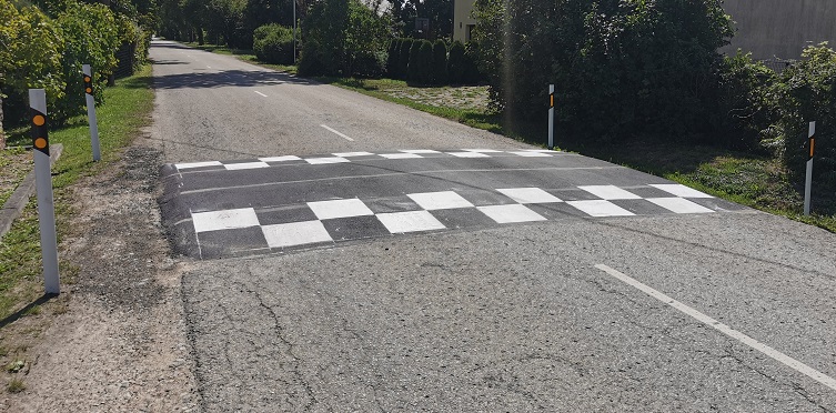 Ceļa ātrumvalnis ar melnbaltu rūtiņu marķējumu uz šauras lauku ielas, abās pusēs izvietoti balti atstarojoši stabiņi un apkārt redzama zaļa veģetācija.