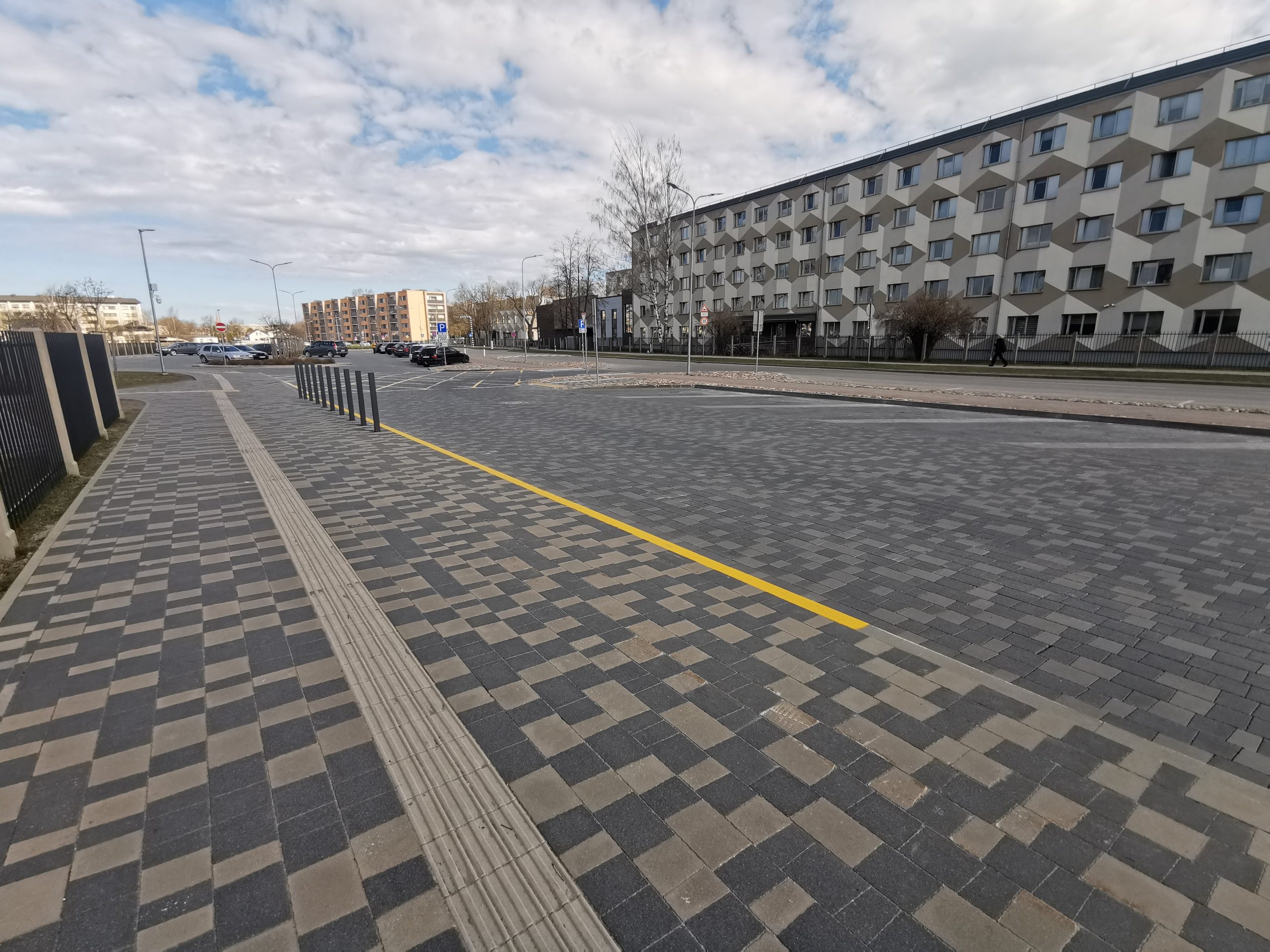 Bruģēts laukums un ietve ar taktilo joslu. Labajā pusē daudzstāvu ēka ar ģeometrisku fasādi, fonā stāvlaukums ar auto un mākoņainas debesis Jelgavā.