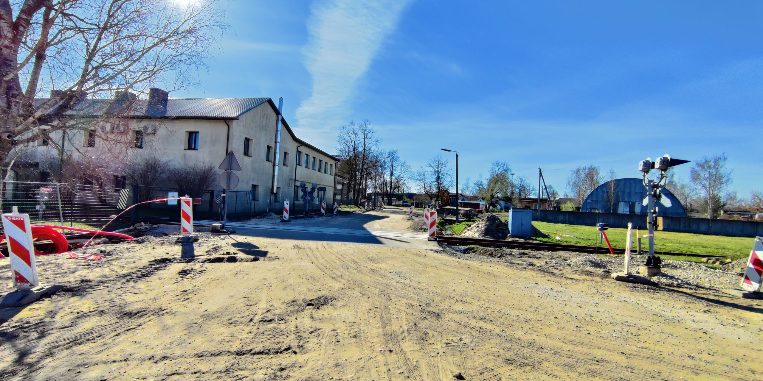 Būvdarbi uz grants ceļa Jelgavā: dzelzceļa pārbrauktuve slēgta, uzstādīti barjeras stabi un zīmes. Fonā divstāvu ēka, blakus bērzs.