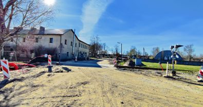 Bauskas_iela Būvdarbi uz grants ceļa Jelgavā: dzelzceļa pārbrauktuve slēgta, uzstādīti barjeras stabi un zīmes. Fonā divstāvu ēka, blakus bērzs.