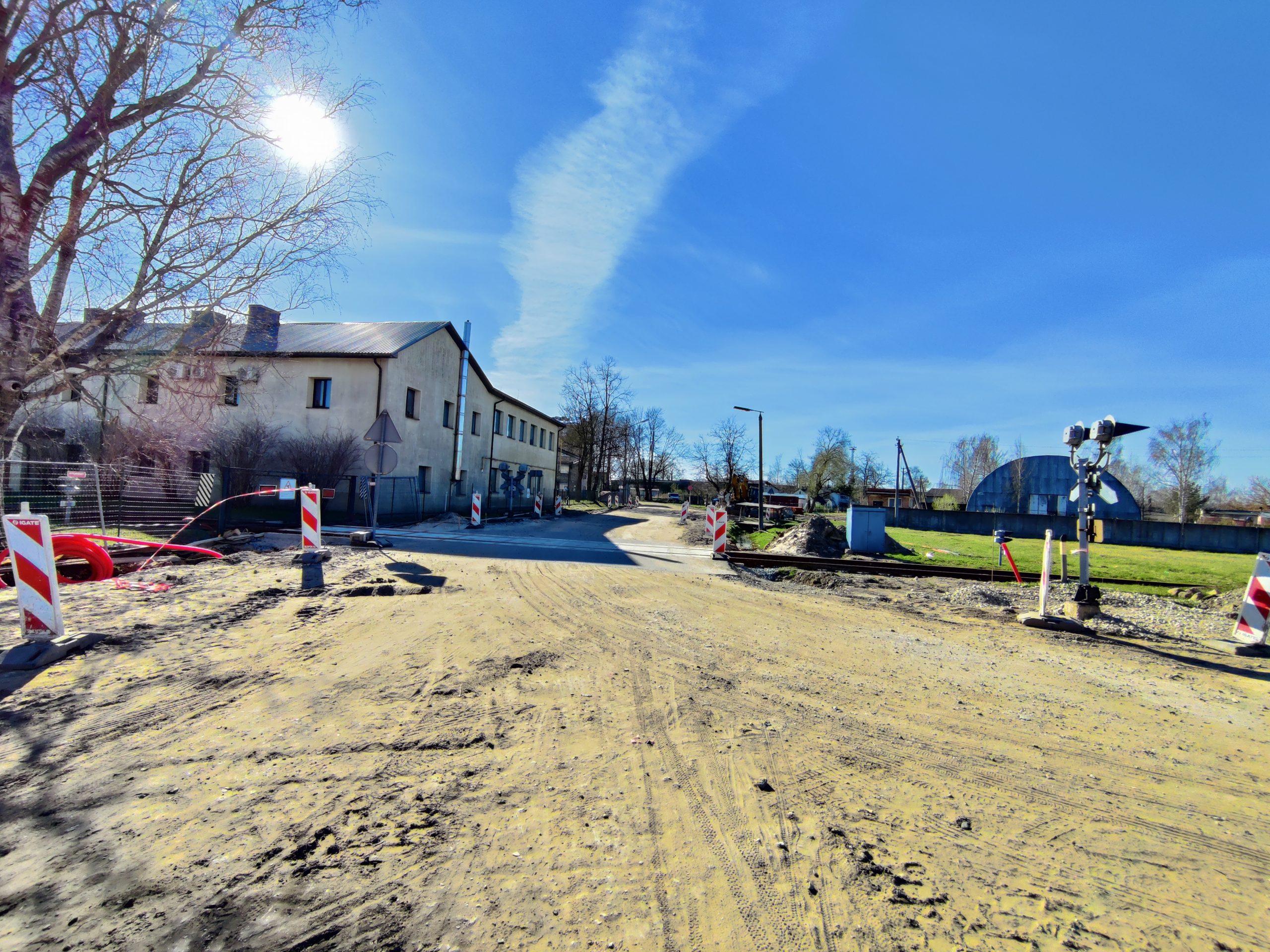 Būvdarbi uz grants ceļa Jelgavā: dzelzceļa pārbrauktuve slēgta, uzstādīti barjeras stabi un zīmes. Fonā divstāvu ēka, blakus bērzs.