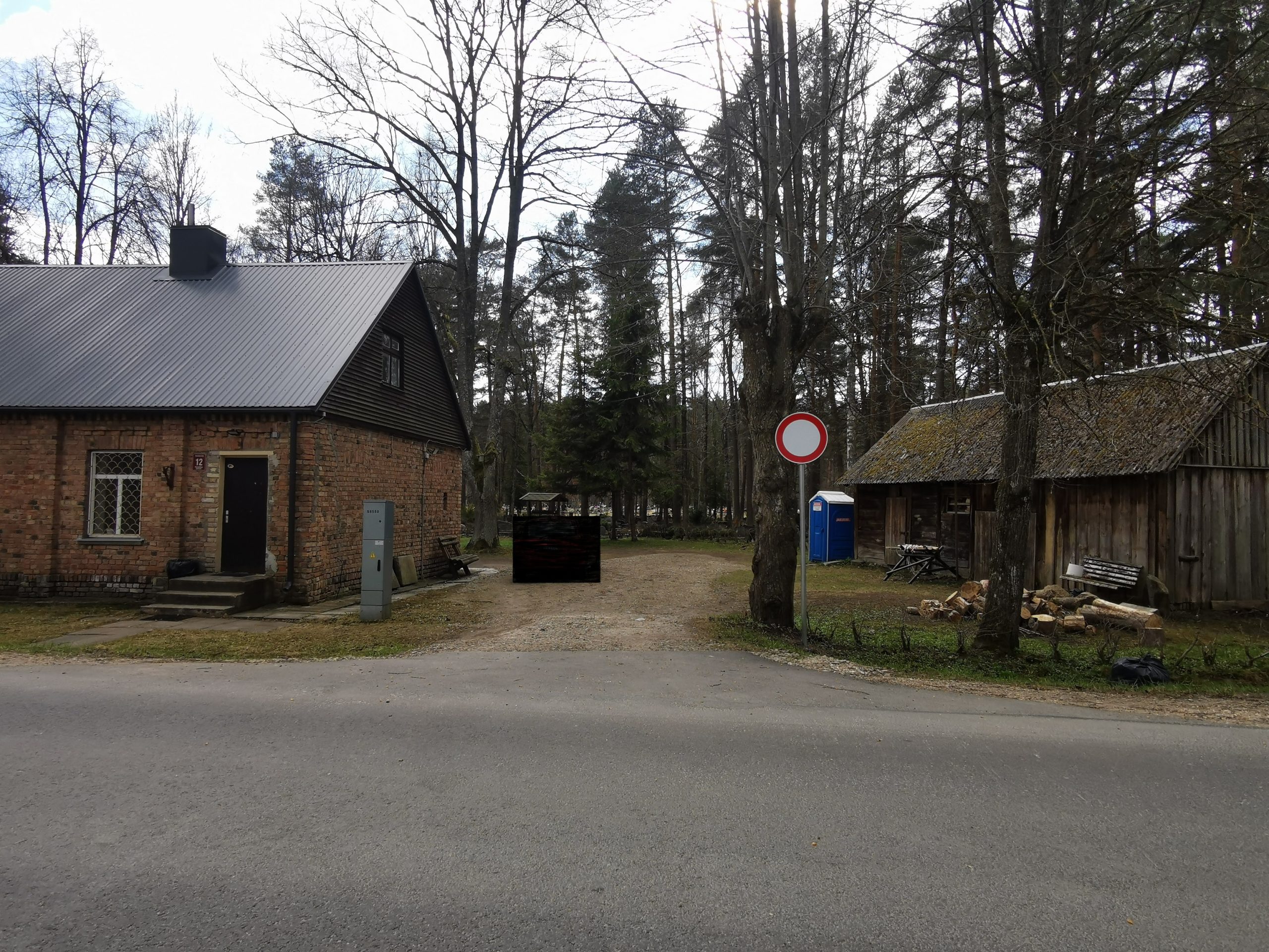 Mūra māja un koka šķūnis meža malā. Starp tiem zemes ceļš ar ceļa zīmi "Braukt aizliegts". Fonā koku ieskauta teritorija.