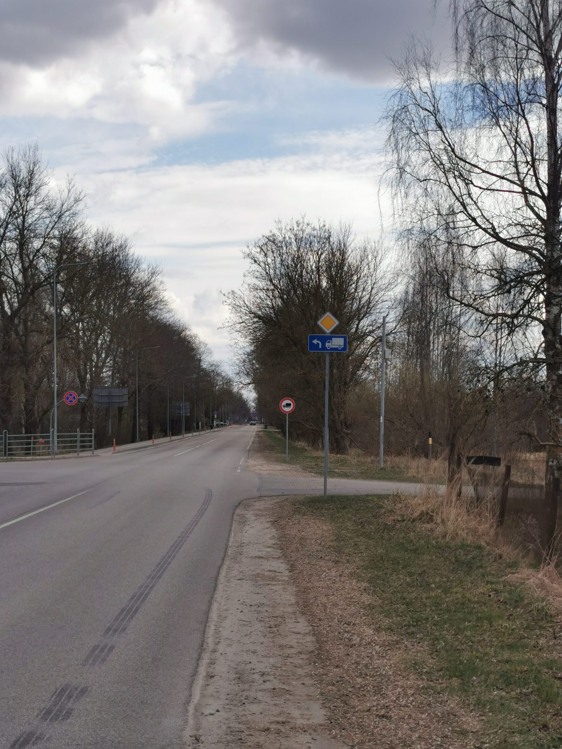 Asfaltēts ceļš ar ceļa zīmēm "Galvenais ceļš" un norādi kravas auto. Malās kaili koki un bērzi, tālumā redzama iela un mākoņainas debesis pavasarī.