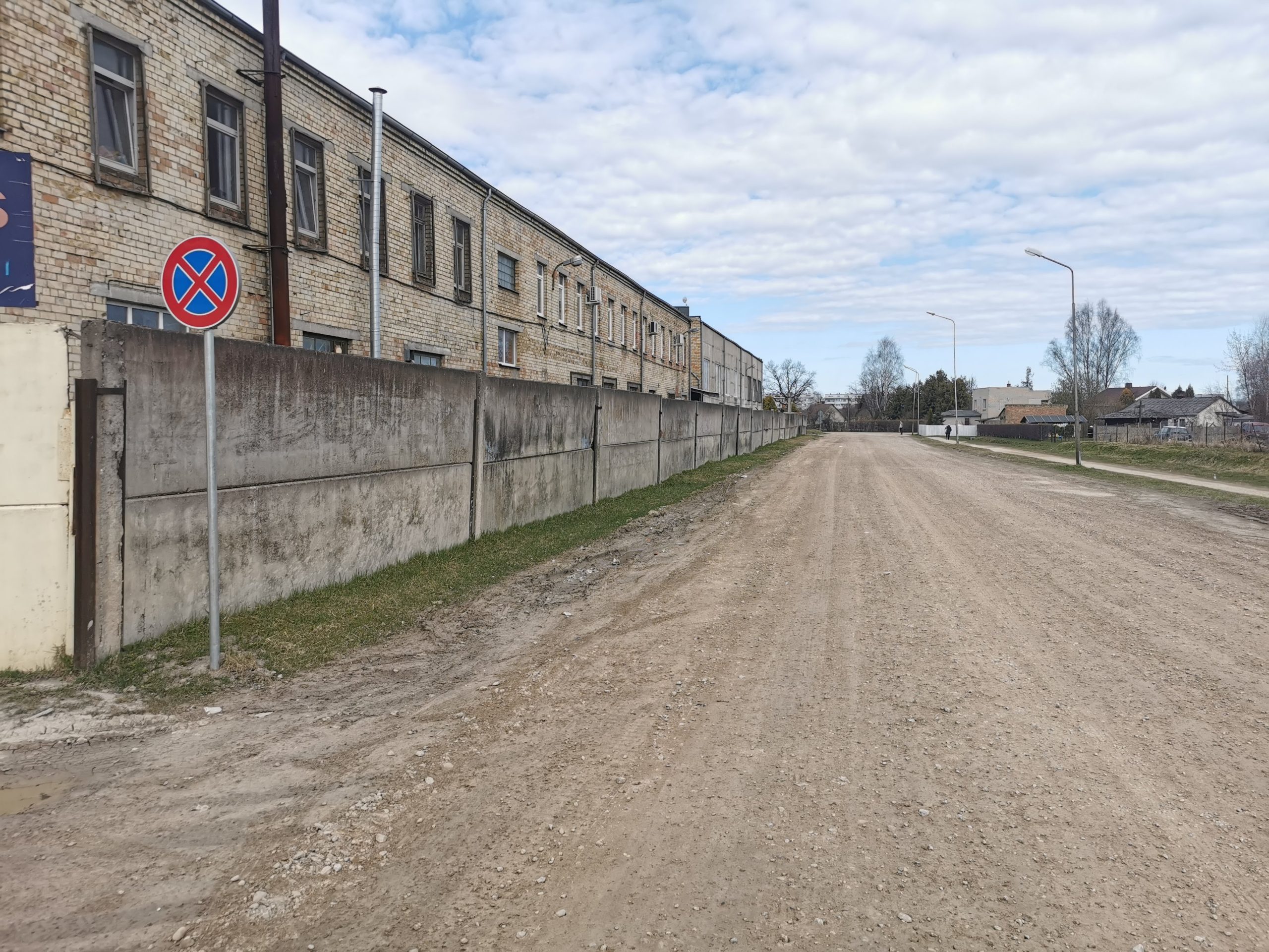 Grants ceļš gar betona žogu un senu mūra ēku. Priekšplānā ceļa zīme "Apstāties un stāvēt aizliegts", fonā ielu apgaismojums un mākoņainas debesis.