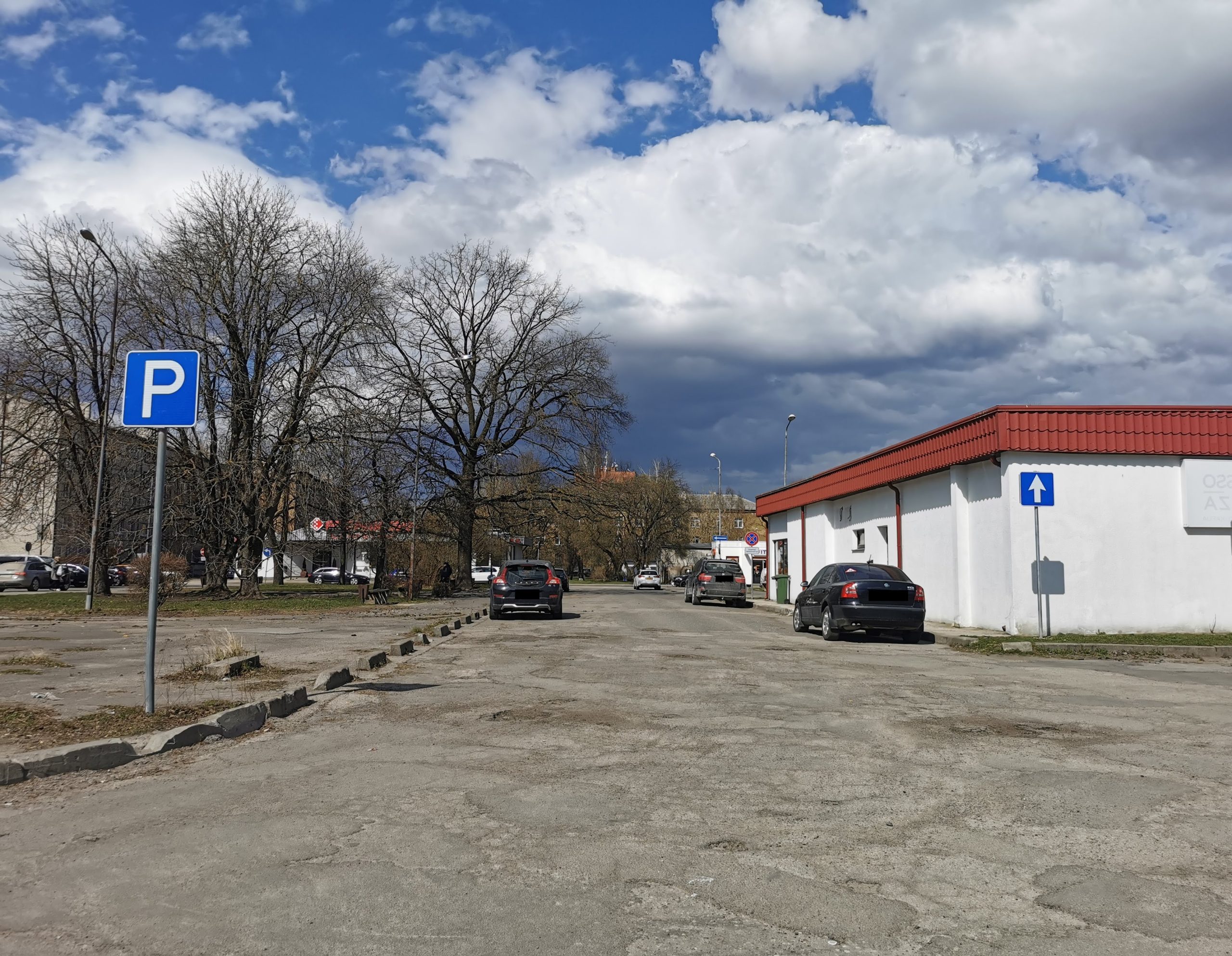 Stāvlaukums ar ceļa zīmi "Stāvvieta". Blakus balta ēka ar sarkanu jumtu, novietotas automašīnas un kaili koki zem zili baltām, mākoņainām debesīm.