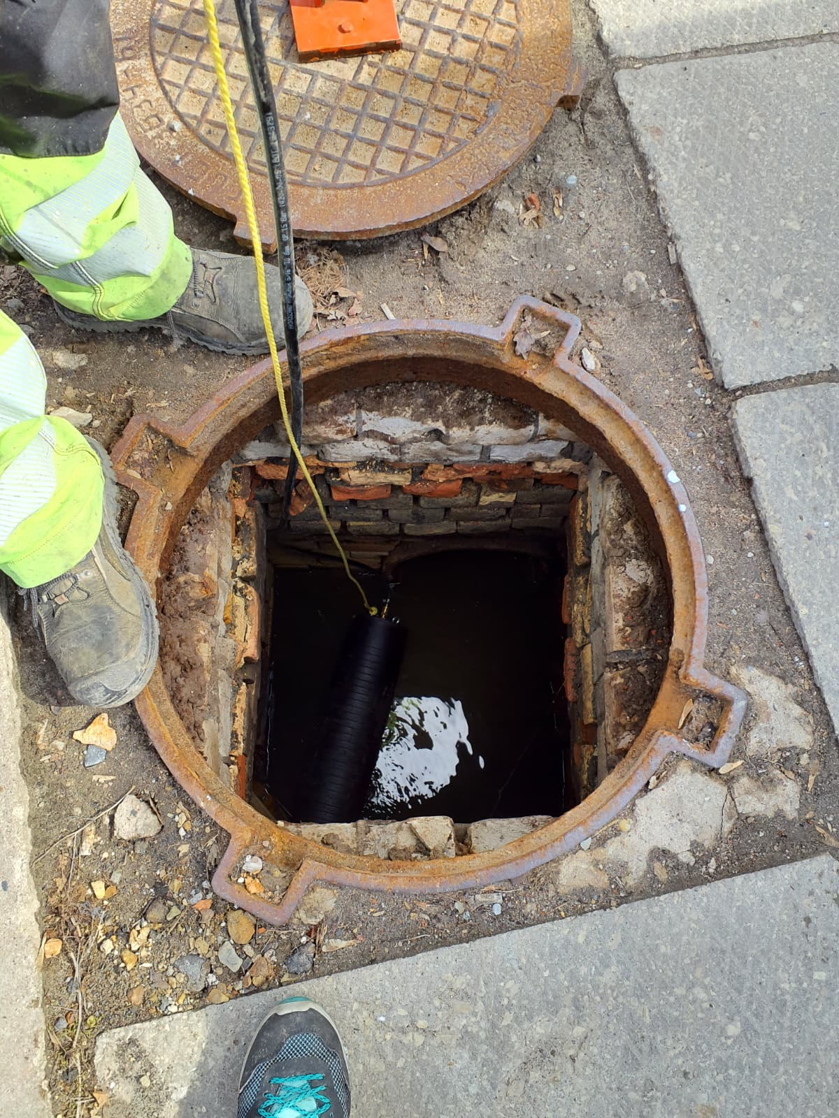 Strādnieks pie atvērtas, mūrētas akas ar ūdeni. Tajā nolaista melna ierīce ar dzeltenu auklu. Blakus uz asfalta guļ noņemts, sarūsējis čuguna vāks.