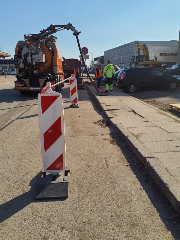 iebrukums_uzvaras (4) Hidrodinamiskā automašīna tīra kanalizāciju. Darba zona norobežota ar svītrainiem vairogiem. Strādnieki uzrauga cauruli, kas nolaista akā.
