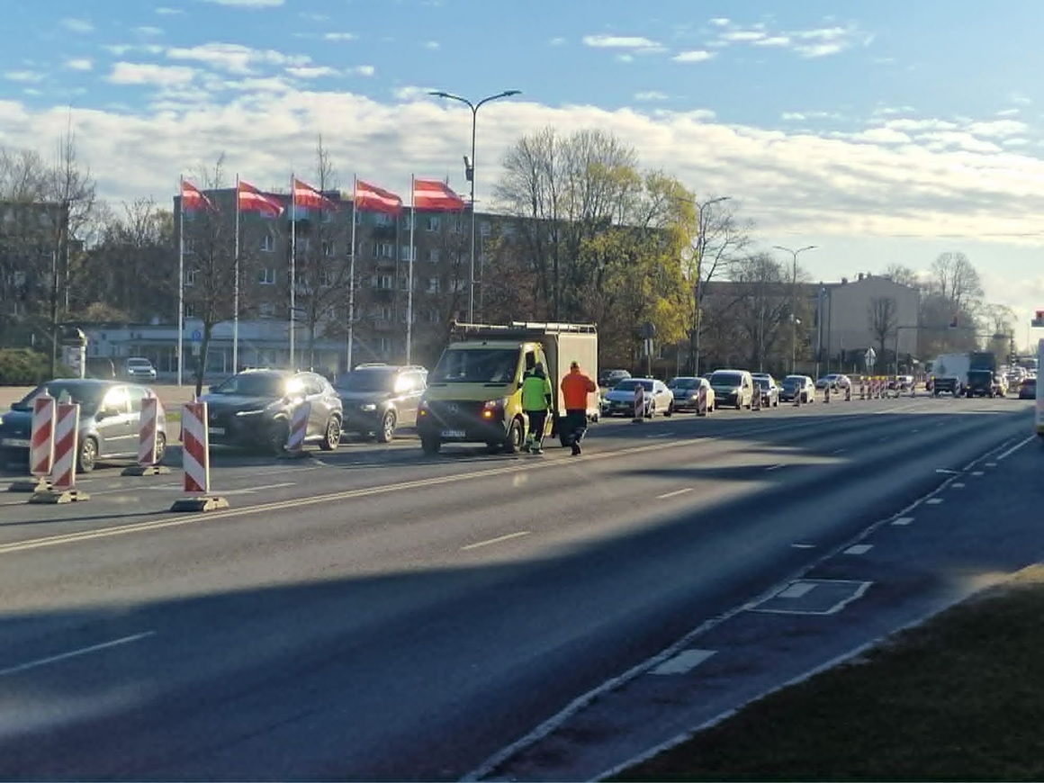 Ceļa remontdarbi Lielajā ielā pie Hercoga Jēkaba laukuma – satiksme vienā joslā, automašīnu rinda un divi ceļstrādnieki pie darba furgona.