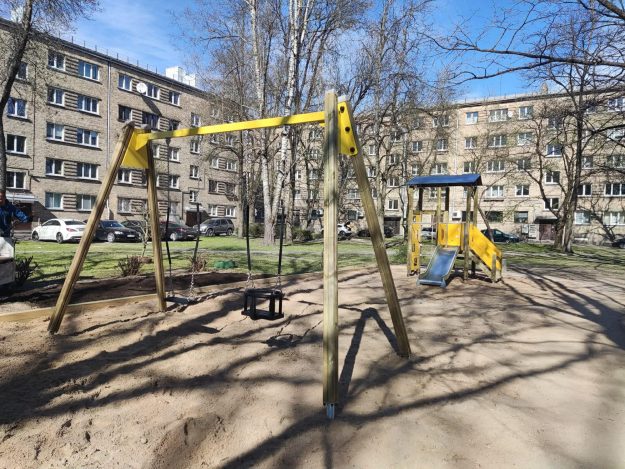 rotalu-laukums-pasta-35b Bērnu rotaļu laukums iekšpagalmā ar koka šūpolēm un slidkalniņu smilšu pamatnē. Fonā redzamas daudzdzīvokļu mājas, koki un automašīnas saulainā dienā.