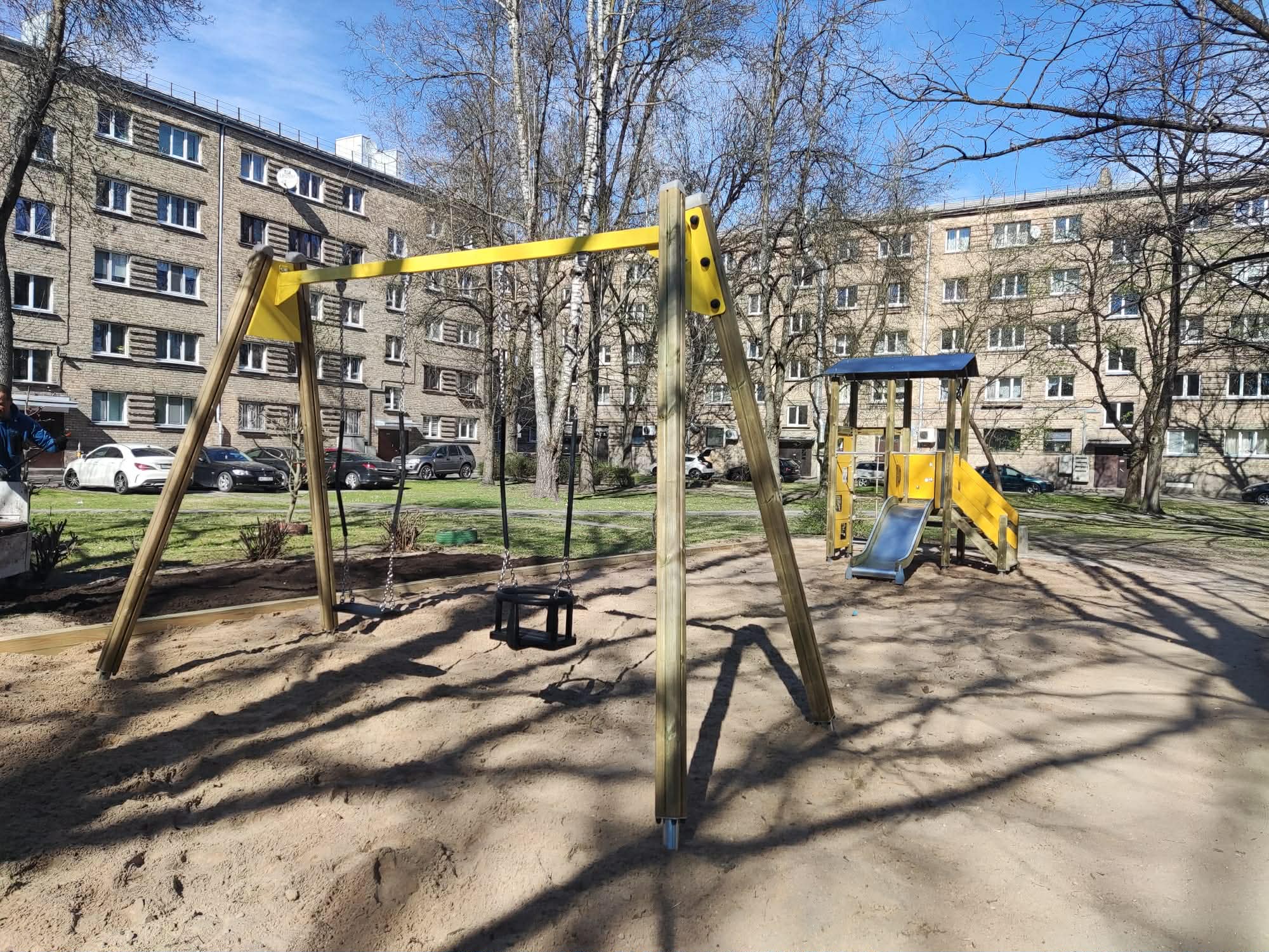 Bērnu rotaļu laukums iekšpagalmā ar koka šūpolēm un slidkalniņu smilšu pamatnē. Fonā redzamas daudzdzīvokļu mājas, koki un automašīnas saulainā dienā.