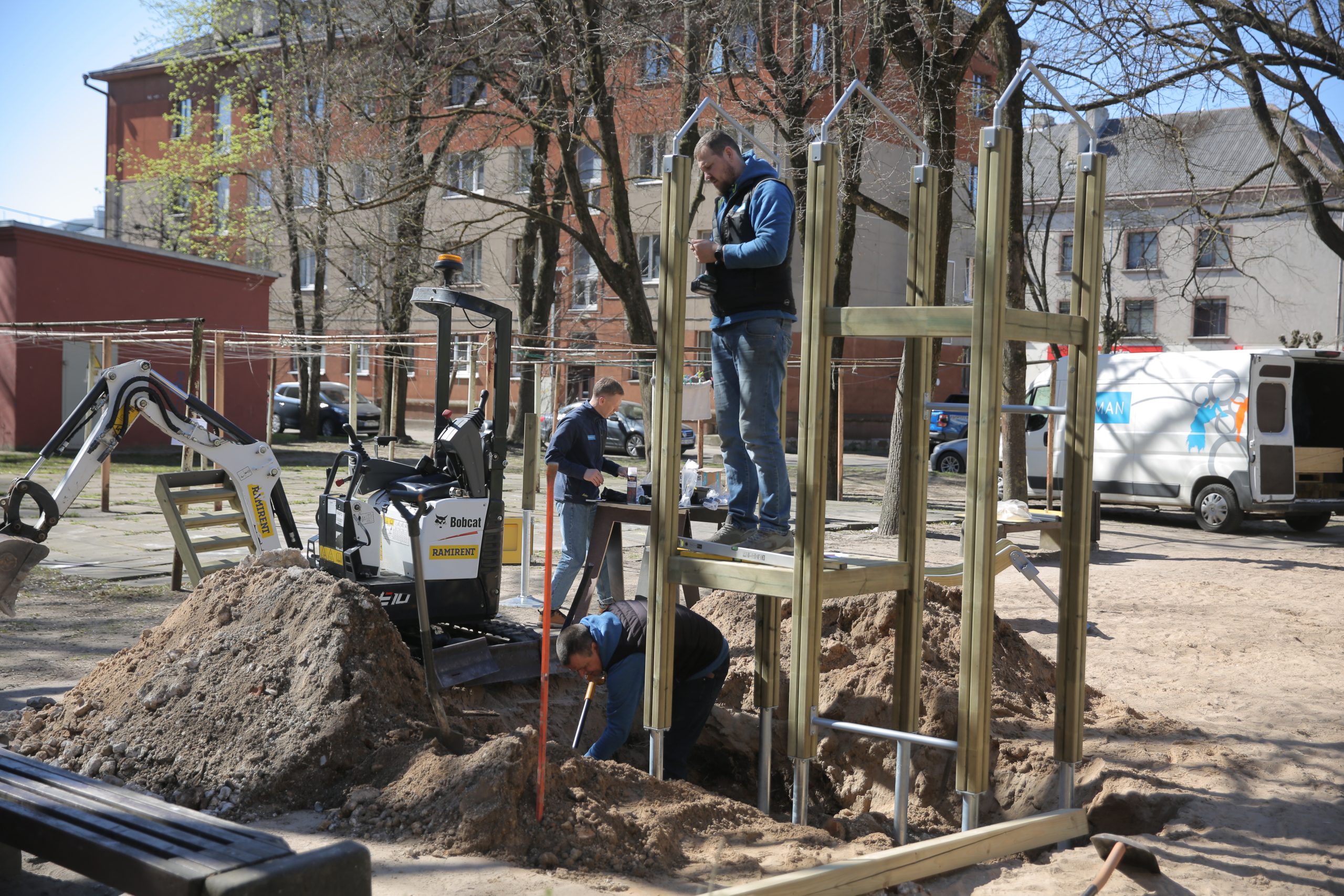 Strādnieki uzstāda koka konstrukciju bērnu rotaļu laukumā. Blakus atrodas mazais ekskavators, smilšu kaudzes un darba instrumenti uz daudzdzīvokļu māju fona.