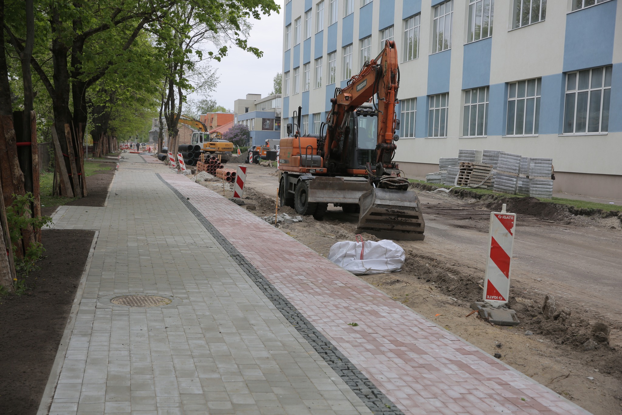 Ielas pārbūve pie ēkas. Ekskavators un strādnieki veic darbus, uzstāda norobežojumus. Ietve no dažādu krāsu bruģa.