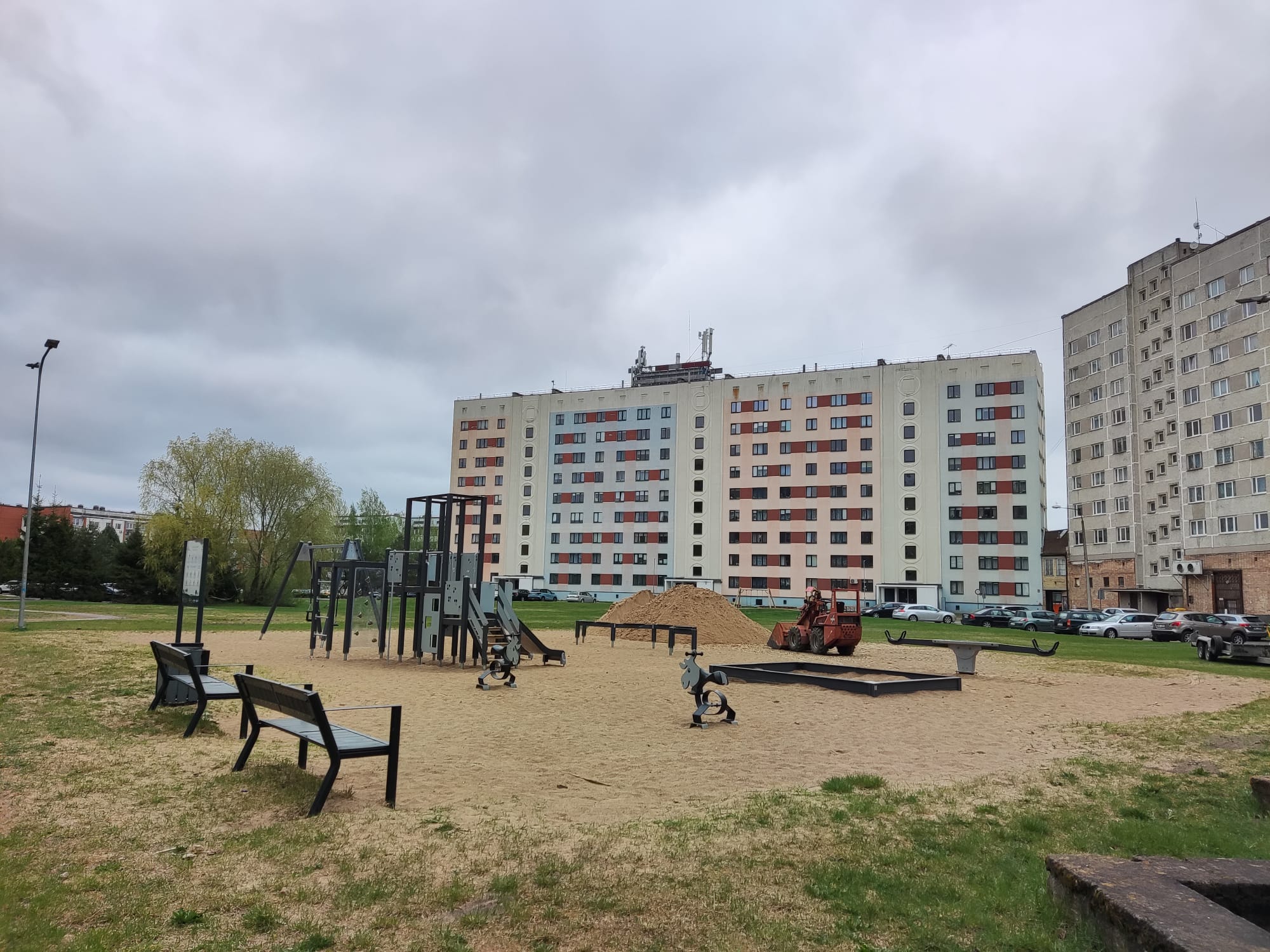 Bērnu rotaļu laukums Jelgavā. Smilšu laukums ar šūpolēm, slidkalniņu, soliņiem. Fonā daudzstāvu dzīvojamā ēka.