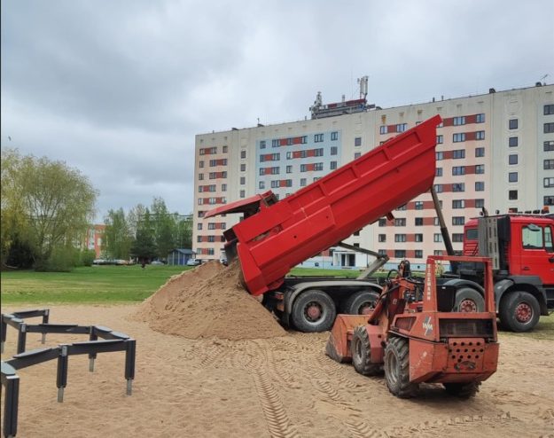 rotalu_laukumi Sarkana kravas automašīna izber smiltis bērnu rotaļu laukumā. Blakus atrodas mazs sarkans traktors un daudzstāvu māja.