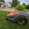 Robots-zāles pļāvējs pļauj zālāju pilsētas parkā. Fonā redzams bērnu rotaļu laukums un koki.