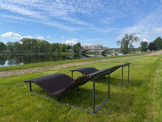 zvilni Atpūtas krēsli zālājā pie Lielupes Jelgavā. Fonā redzama upe, koki un Mītavas tilts zem skaidrām debesīm.