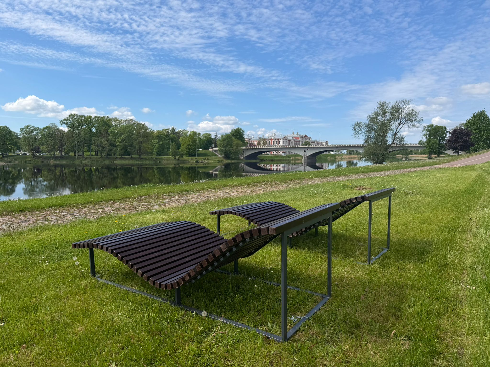 Atpūtas krēsli zālājā pie Lielupes Jelgavā. Fonā redzama upe, koki un Mītavas tilts zem skaidrām debesīm.