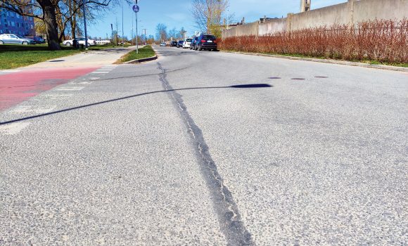 Neretas ielas un Birzes ielas krustojuma asfaltā redzama gareniska plaisa; ceļa malā novietotas automašīnas