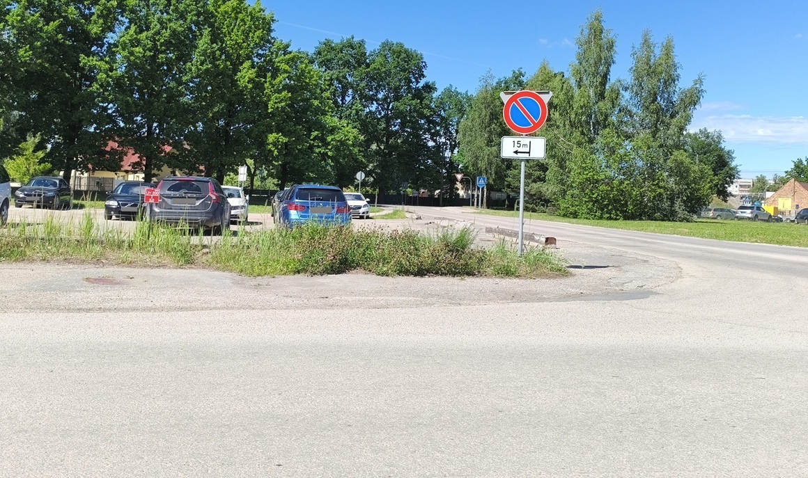 Ceļa zīme “Apstāties aizliegts” ar papildzīmi, kas norāda aizlieguma sākumu pēc 15 metriem pa kreisi. Fonā redzama autostāvvieta, koki un ceļš.