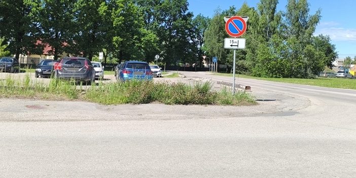 Ceļa zīme “Apstāties aizliegts” ar papildzīmi, kas norāda aizlieguma sākumu pēc 15 metriem pa kreisi. Fonā redzama autostāvvieta, koki un ceļš.