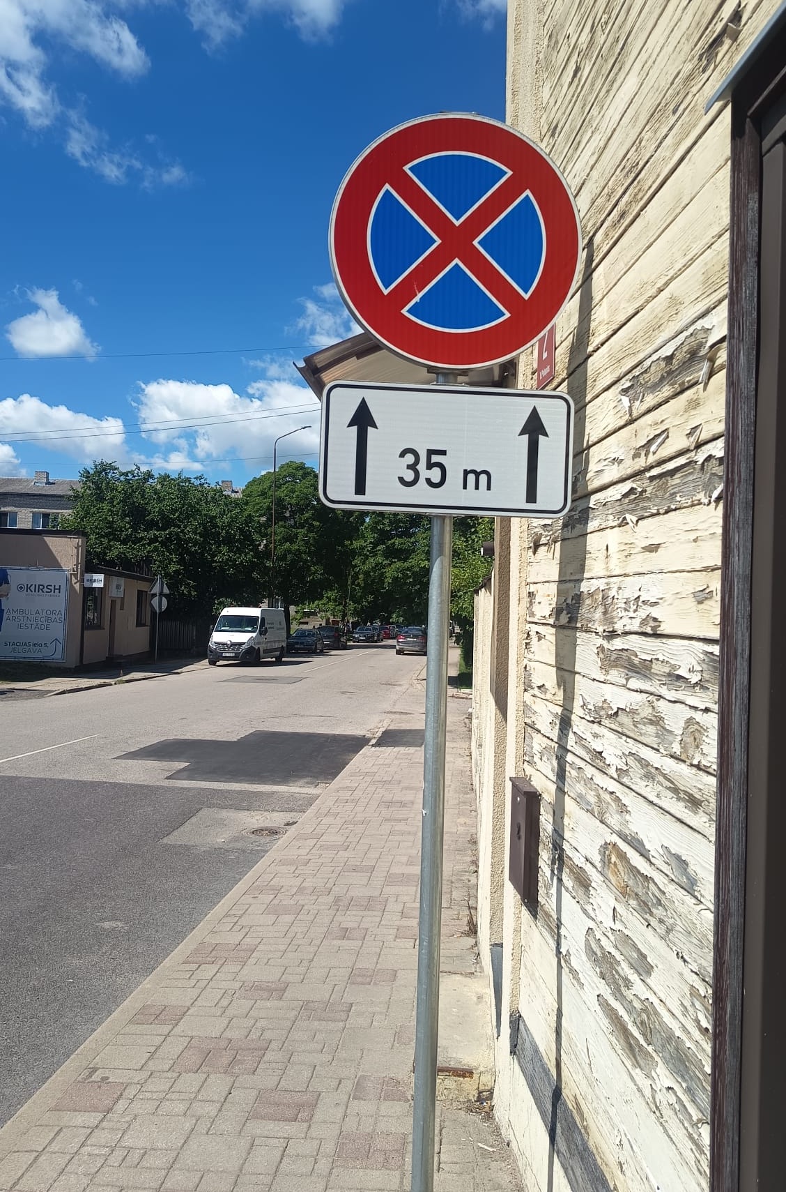 Ceļa zīme “Apstāties aizliegts” ar papildzīmi, kas norāda aizlieguma zonu 35 metru garumā uz priekšu. Zīme uzstādīta pie ēkas gar ielu.