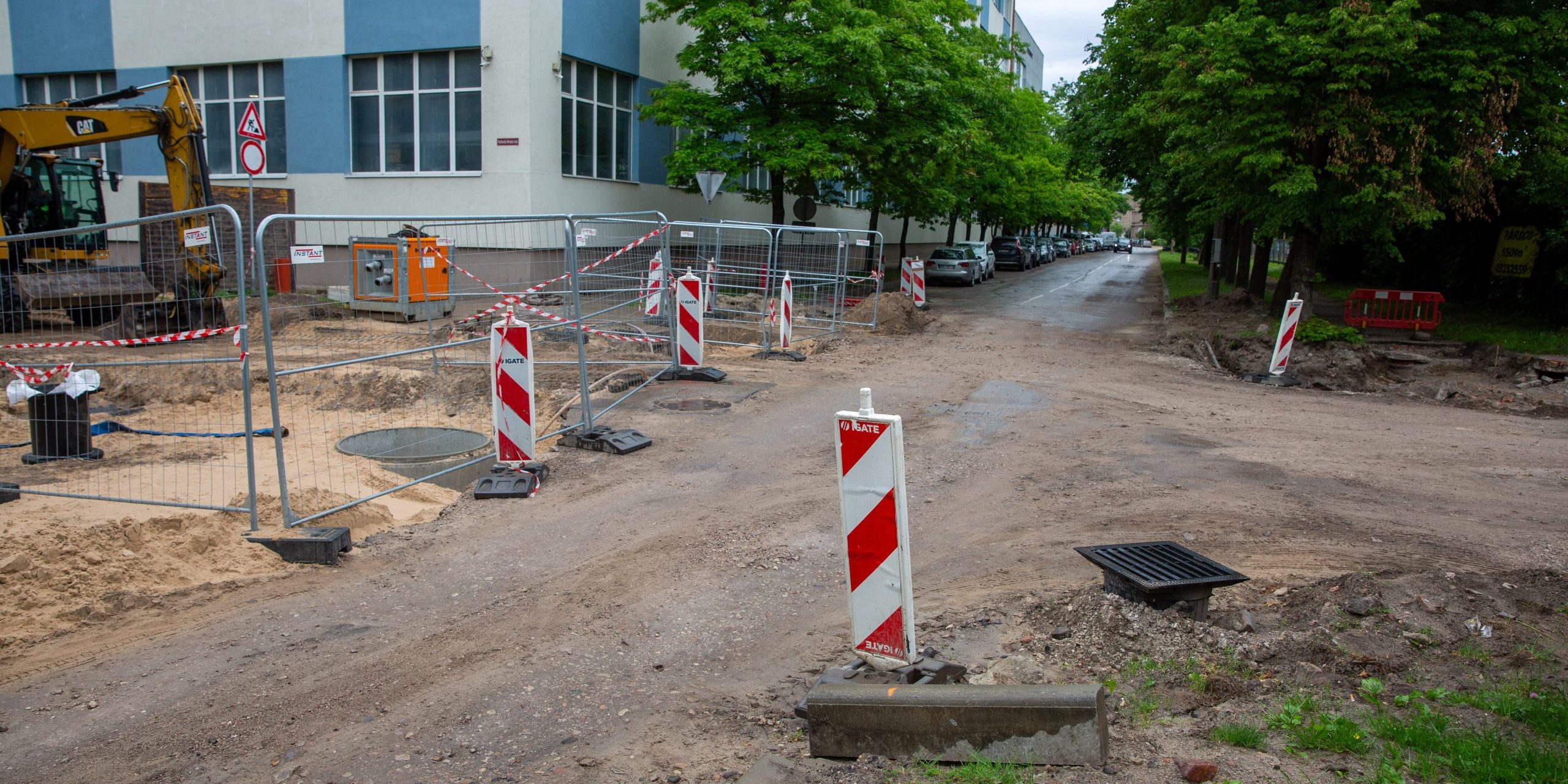 Ielas krustojuma remontdarbi pilsētvidē ar norobežojošām metāla sētām, vadstatņiem un ekskavatoru fona.
