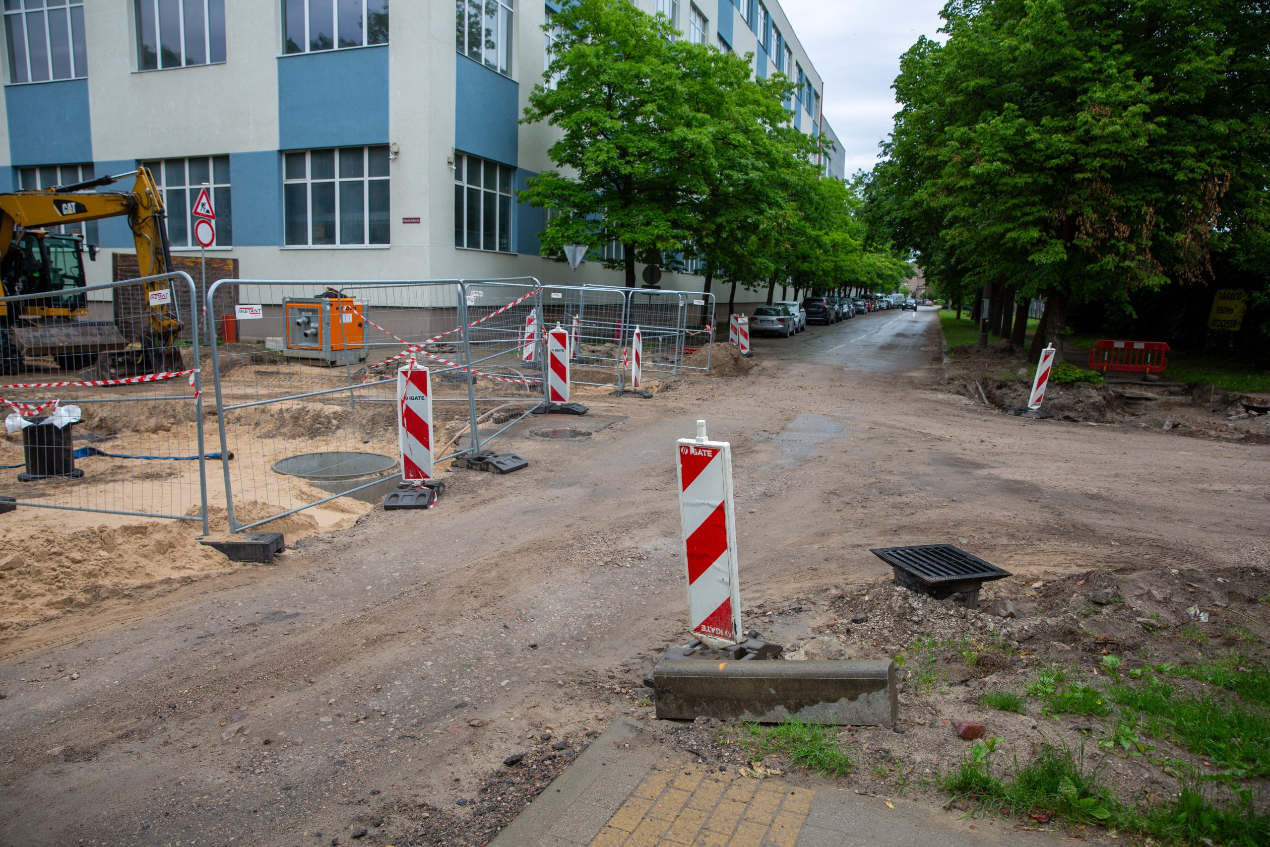 Ielas krustojuma remontdarbi pilsētvidē ar norobežojošām metāla sētām, vadstatņiem un ekskavatoru fona.