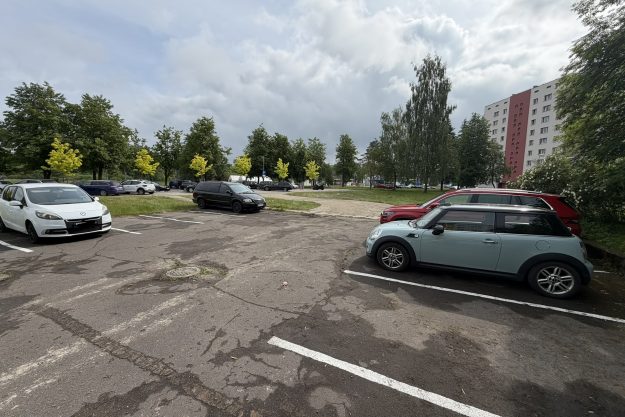stavvieta_loka_magistrale Automašīnu stāvlaukums pie daudzdzīvokļu mājas ar zaļu pagalmu un kokiem fona.