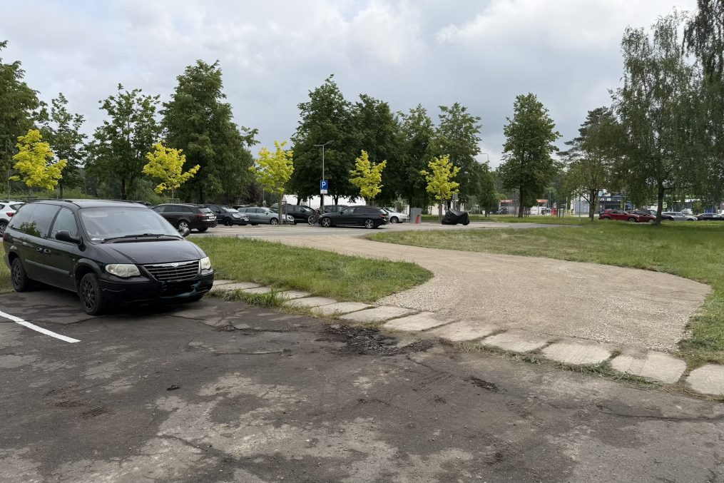 Automašīnu stāvlaukums pie daudzdzīvokļu mājas ar zaļu pagalmu un kokiem fona.