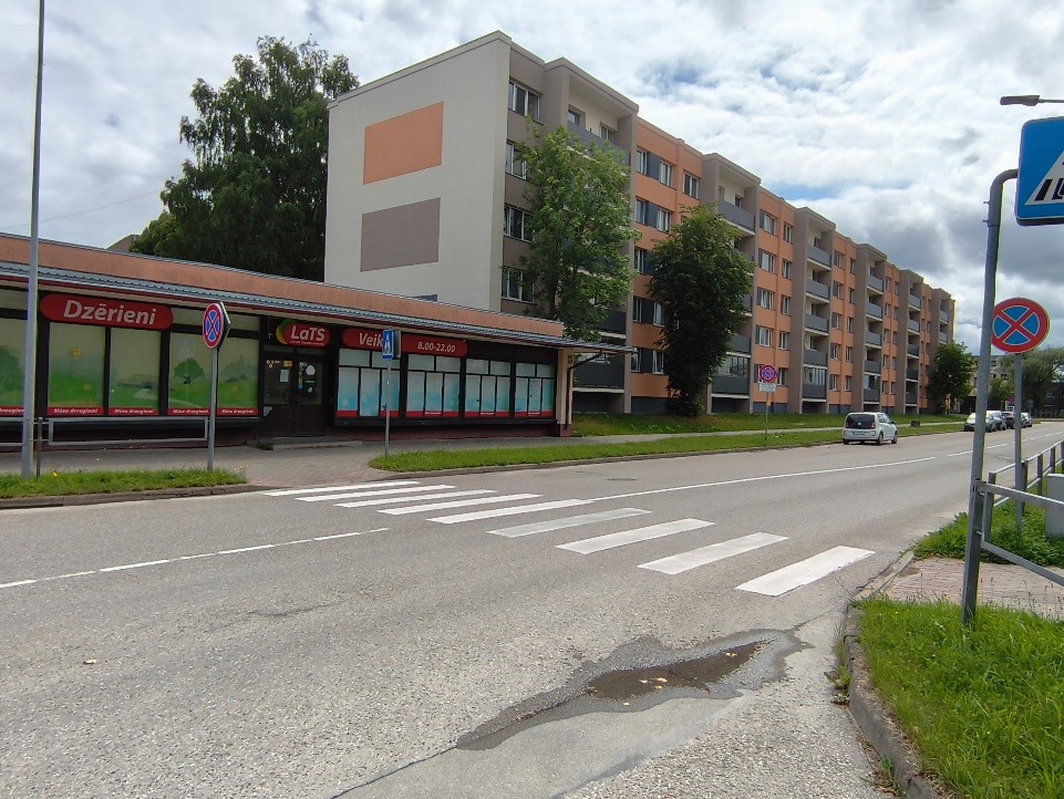 Pulkveža Oskara Kalpaka iela ar gājēju pāreju, daudzdzīvokļu dzīvojamo ēku. Gar ielu novietotas ceļa zīmes, fonā koki un mākoņainas debesis.