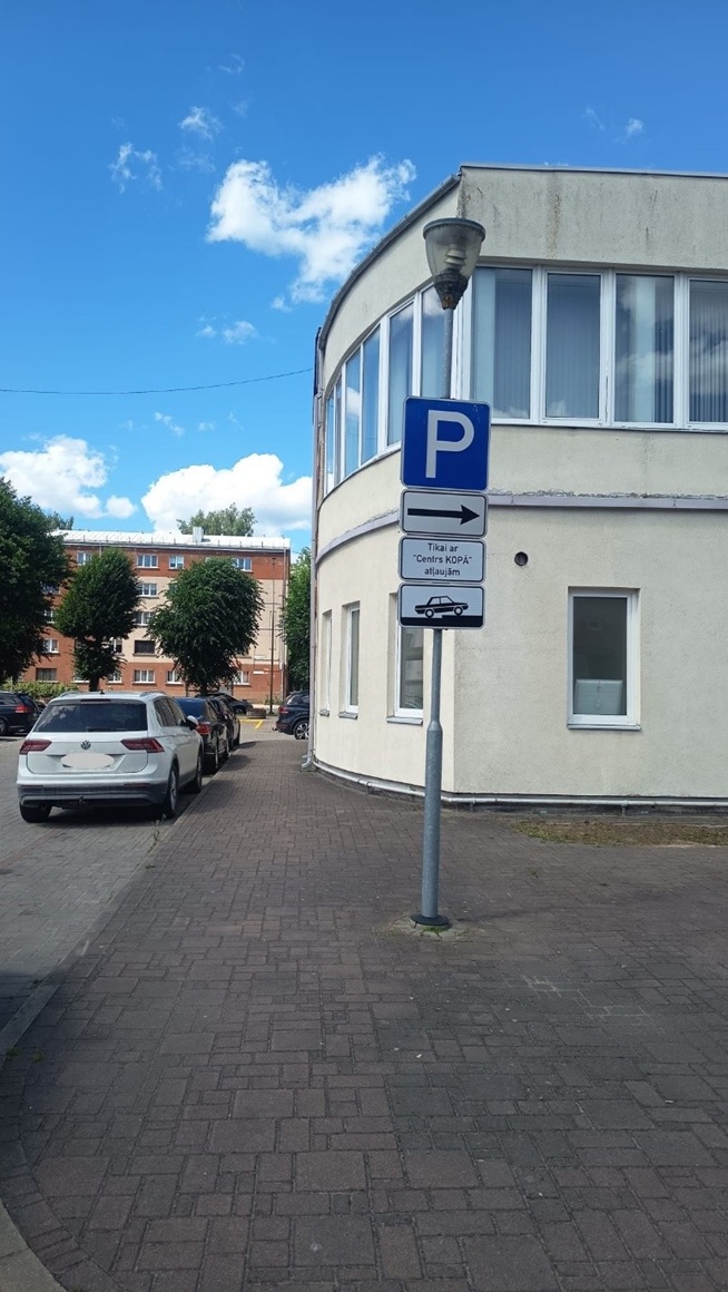 Pie ēkas uz ietves redzama autostāvvietas zīme ar norādi pa labi. Papildzīmes informē, ka stāvvieta paredzēta tikai “Centrs KOPA” atļaujām. Kreisajā pusē novietotas automašīnas.