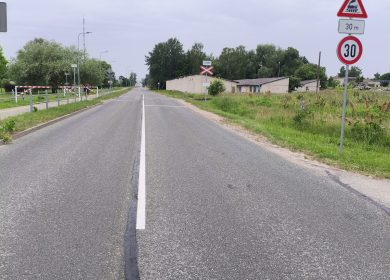 Prohorova Prohorova iela pie dzelzceļa pārbrauktuves. Redzamas brīdinājuma zīmes, 30 km/h ātruma ierobežojums, pārbrauktuves barjeras, ceļmala ar zāli un ēkas.