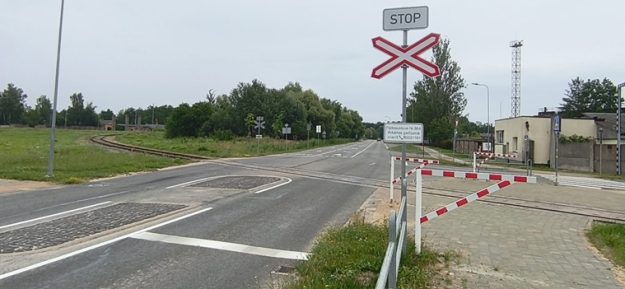 Prohorova_Neretas_small Prohorova iela pie dzelzceļa pārbrauktuves. Redzamas brīdinājuma zīmes, 30 km/h ātruma ierobežojums, pārbrauktuves barjeras, ceļmala ar zāli un ēkas.