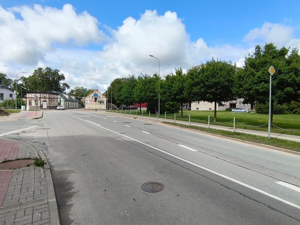 Raiņa ielas un Jēkaba ielas krustojums, redzams luksofors un zaļā zona gar brauktuvi. Ielas malās aug koki, redzamas dzīvojamās ēkas.