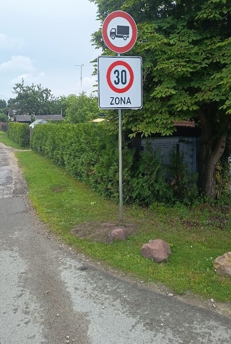 Ruļļu iela ar ceļa zīmi, asfaltēts ceļš. Labajā pusē redzama ceļa zīme "Maksimālā ātruma ierobežojuma zona 30" un “Kravas automobiļiem braukt aizliegts”, zaļā zona un dzīvžogs.