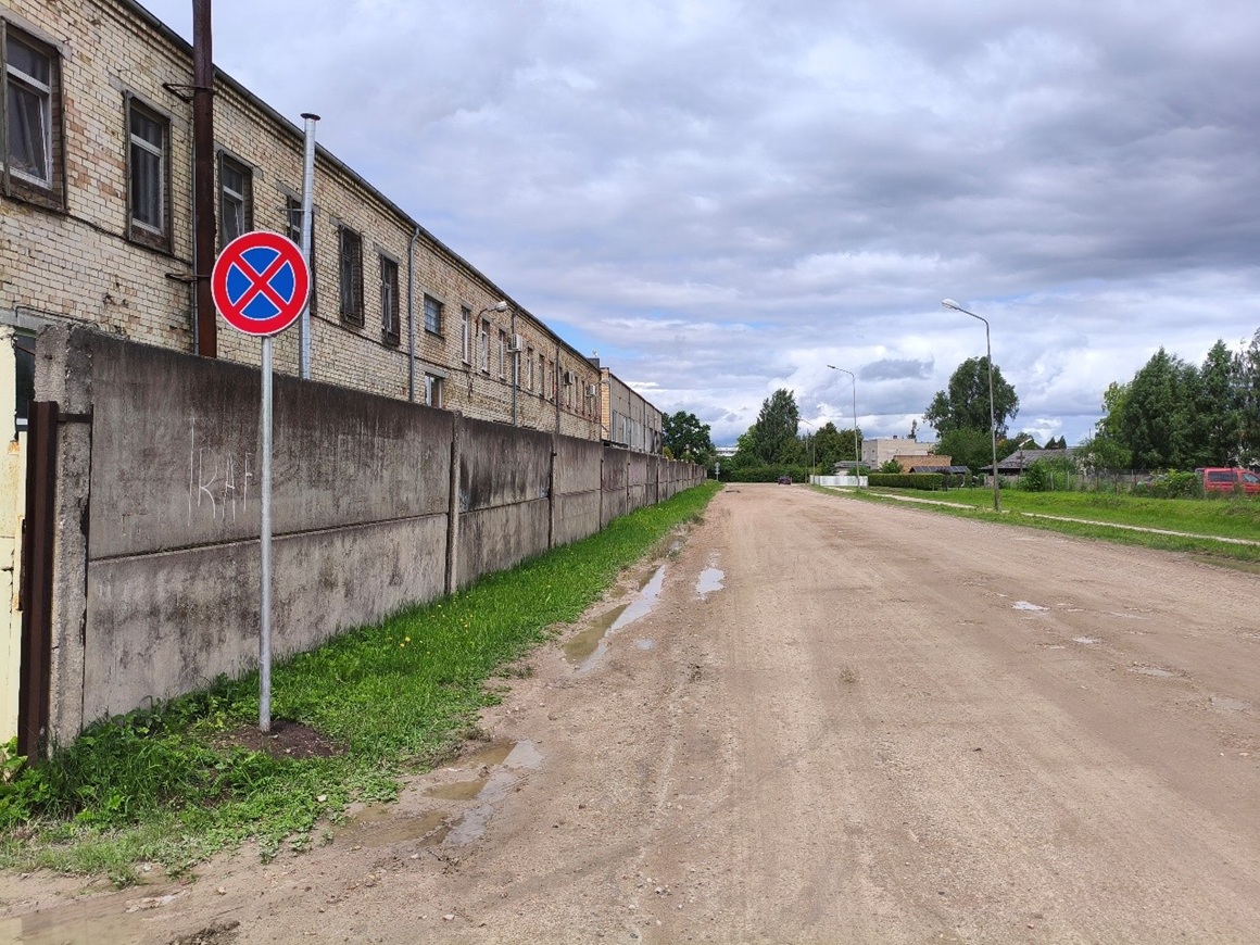 Ceļš ar zaļo zonu gar malām, kreisajā pusē ēka ar betona žogu un ceļa zīme "Apstāties aizliegts".
