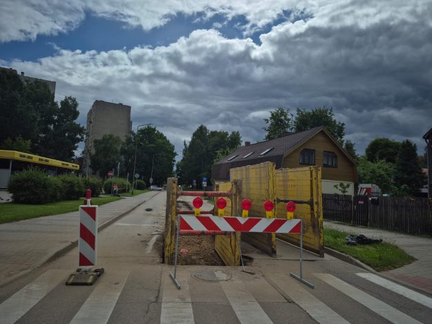 Māras iela Cel̦a remontdarbi pilsētā ar barjerām, dzeltenām sienām un brīdinājuma gaismām. Tālumā redzamas mājas un mākoņainas debesis.