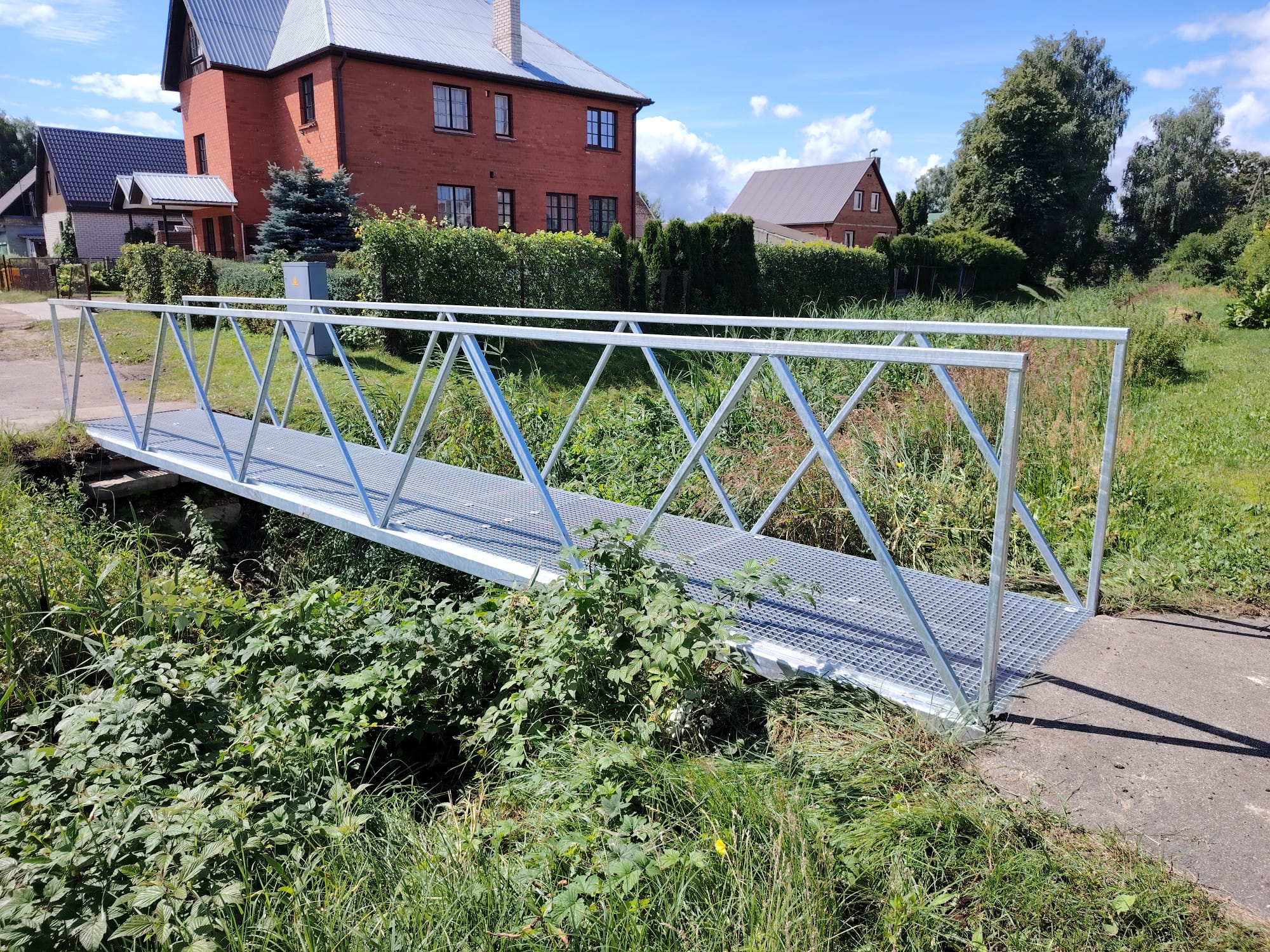 Jauna cinkota metāla gājēju tiltiņa fotogrāfija pār nelielu grāvi, savienojot asfalta celiņu un zālienu.
