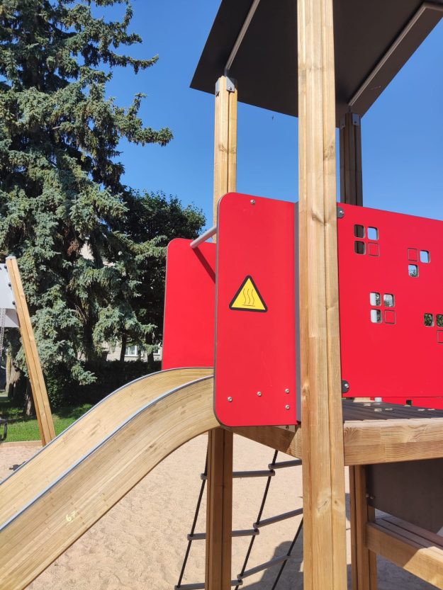 slidkalnini (1) Koka rotaļu laukuma konstrukcija ar sarkanu paneli, slidkalniņu un dzeltenu brīdinājuma zīmi "Karsta virsma"